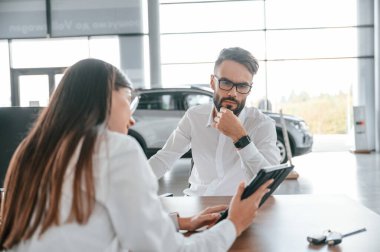 Tablette bilgi gösteriliyor. Masanın yanında oturuyor. Beyaz elbiseli bir adam araba galerisinde..