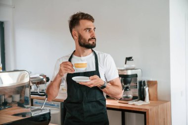 Ayakta ve bardağı tutarken. Beyaz gömlekli ve siyah önlüklü kafe çalışanı içeride..