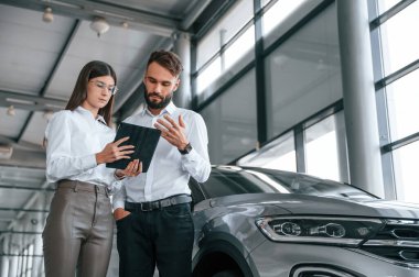 Siyah tablet ellerde. Bilgi okunuyor. Beyaz elbiseli bir adam araba galerisinde birlikte..