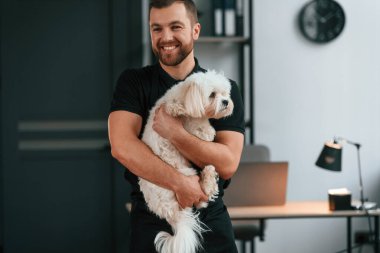 Adam gülümsüyor. Groomer içeride Maltalı köpekle birlikte. Siyah üniformalı adam..