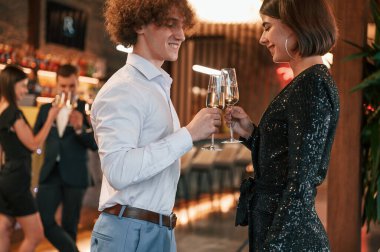 Kadın ve erkek ayakta duruyor ve poz veriyor. Güzel giyinmiş bir grup insan içeride yeni yılı kutluyorlar..