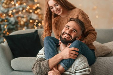 Kadın kanepede, erkeğini bacaklarından kucaklıyor. Güzel genç çift yeni yılı evde kutluyor..