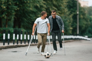 Futbol topuyla oynamak. Koltuk değnekli iki adam yolda..