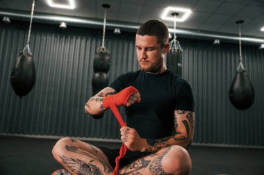 Oturuyor ve kırmızı el sargıları takıyor. Dövmeli genç adam spor salonunda boks antrenmanı yapıyor..