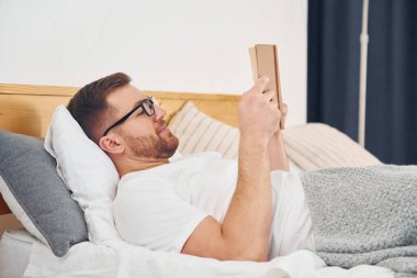 Beyaz gömlekli ve gözlüklü bir adam yatağa uzanıp kitap okuyor..