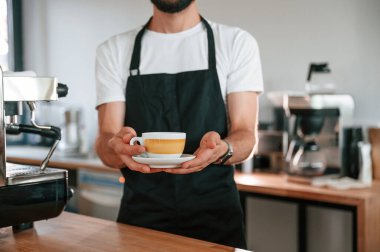 Kupa ellerde. Beyaz gömlekli ve siyah önlüklü kafe çalışanı içeride..