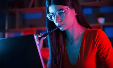 Stylus 'u tutuyorum. Gözlüklü ve kırmızı elbiseli güzel bir kadın karanlık odada dizüstü bilgisayarının yanında oturuyor..