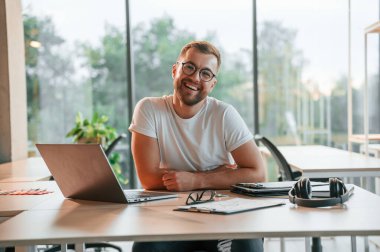 Oturuyor ve gülümsüyor. Modern ofiste resmi giyinmiş olumlu bir adam çalışıyor..