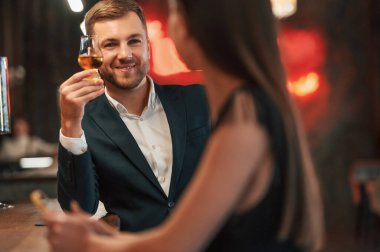 Gülümsüyor, birbirleriyle konuşuyorlar. Kadın ve erkek barda oturmuş içki içiyorlar..
