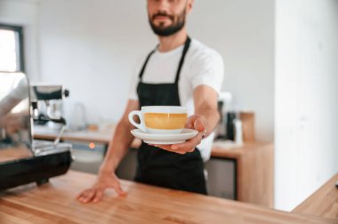 Ayakta ve bardağı tutarken. Beyaz gömlekli ve siyah önlüklü kafe çalışanı içeride..