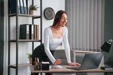 Dizüstü bilgisayarla. Ayakta ve çalışıyor. Beyaz elbiseli genç ve güzel bayan ofiste..