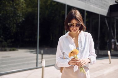 Büyük pencereli binaya karşı. Güzel esmer kadın elinde çiçeklerle dışarıda dikiliyor..