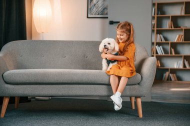 Sarı elbiseli küçük kız kanepede oturuyor ve evde köpeğiyle oturuyor..