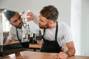 Kahve makinesini temizliyorum. Beyaz gömlekli ve siyah önlüklü kafe çalışanı içeride..
