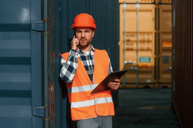 Telefon kullanarak ve tablet tutarak. Erkek işçi konteynırlarla birlikte olay yerinde..