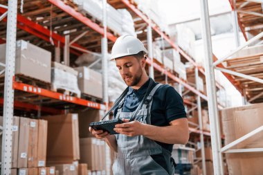 Beyaz kasketli ve tabletli. Üniformalı eleman gündüz vakti depoda çalışıyor..