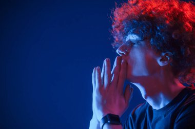 Yan görüş. Kıvırcık saçlı genç adam kapalı alanda neon ışıkla aydınlatılır..