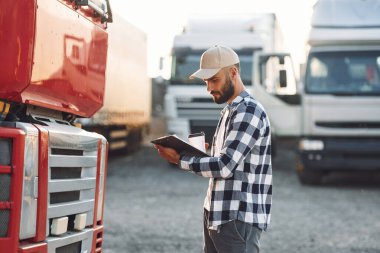 With black notepad. Young truck driver is with his vehicle at daytime. clipart