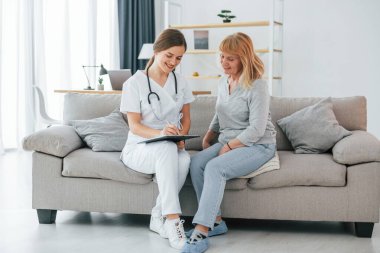 Hemşire reçete yazıyor. Üst düzey bir kadın, içeride genç bir bayan doktorla görüşüyor..