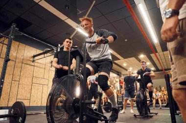 Süreci elinde not defteri olan antrenör kontrol ediyor. Bisiklette kardiyo egzersizi yapan sporcu..