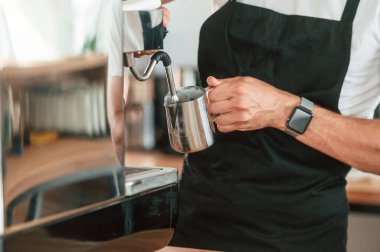 Kahve makinesiyle çalışmakla meşgul. Beyaz gömlekli ve siyah önlüklü kafe çalışanı içeride..