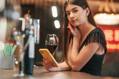 Genç bir kadın bir bardak içkiyle barda oturuyor ve elinde akıllı telefon var..