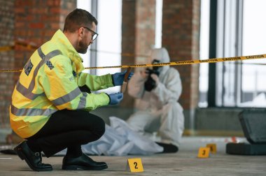 Kadın fotoğraf makinesi kullanıyor, fotoğraf çekiyor. Dedektifler cesedin yanındaki suç mahallinde kanıt topluyorlar..