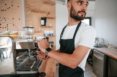 Kahve makinesini kullanarak. Beyaz gömlekli ve siyah önlüklü kafe çalışanı içeride..