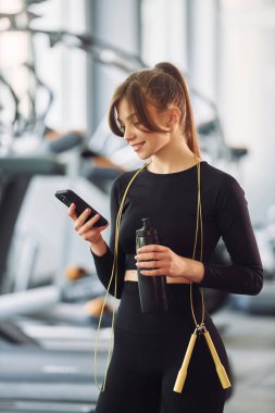 Ellerinde telefon ve boynunda ip ile. Spor elbiseli genç bir kadın spor salonunda spor yapıyor..