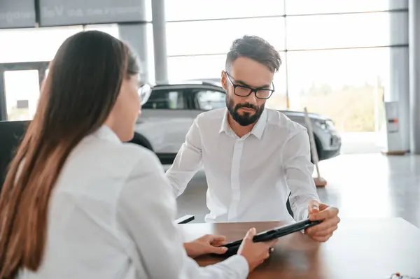 stock image Showing information in the tablet. Sitting by the table. Man with woman in white clothes are in the car dealership.