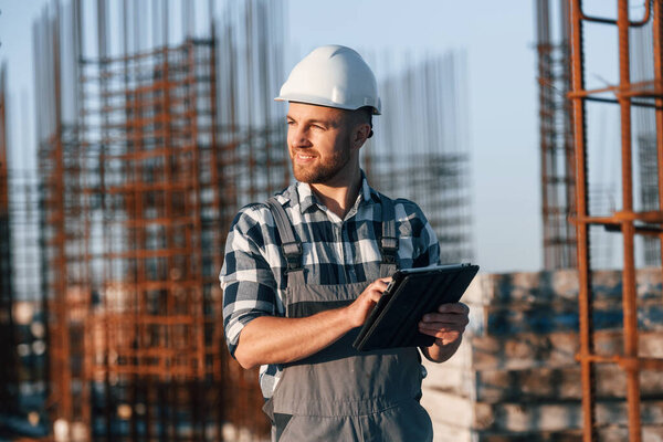 Looking to the side. Holding tablet. Man is working on the construction site at daytime.