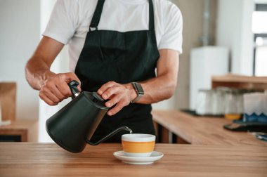 Kahve hazır. Malı bardağa döküyorum. Beyaz gömlekli ve siyah önlüklü kafe çalışanı içeride..