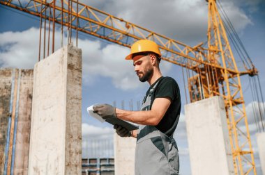 Sarı vince karşı durmak ve elinde belgelerle not defteri tutmak. Üniformalı adam inşaat alanında çalışıyor..