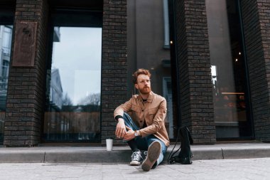 Kahve fincanı. Sakallı, haki renkli ceketli ve kot pantolonlu şık bir adam binanın yanında..