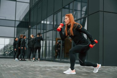 Dambıllarla egzersiz yapıyorum. Bir grup sportif kadın dışarıda, siyah binanın yakınında..