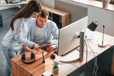 Adam oturuyor, kadın ayakta. Bilgisayarı kullanarak. İki genç tasarımcı ofiste birlikte çalışıyor..