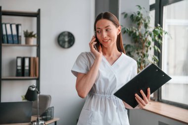 Ayakta duruyor, telefonla konuşuyor ve not defterini tutuyor. Beyaz elbiseli güzel bir kadın..