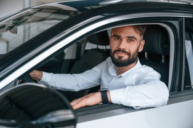 Oturmuş, otomobilin camından bakıyor. Beyaz elbiseli genç adam araba galerisinde..