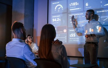 İnsanlar oturuyor. Erkek lider çalışanlarla konuşuyor. Borsa şirketinin projektörüyle ilgili planı gösteriyor..