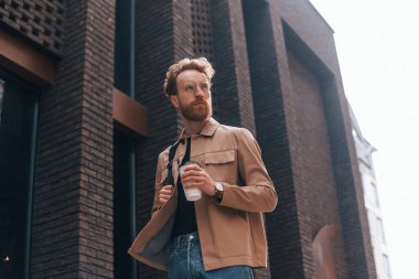 Kahve fincanı. Sakallı, haki renkli ceketli ve kot pantolonlu şık bir adam binanın yanında..