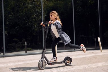 Yan görüş. Küçük kız açık hava binasının yakınında motosiklete biniyor..