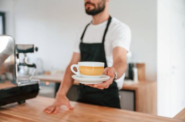 Ayakta ve bardağı tutarken. Beyaz gömlekli ve siyah önlüklü kafe çalışanı içeride..