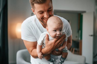 Ellerinde bebekle. Gülümsüyor, eğleniyor. Bebekli baba evde, oğluyla ilgileniyor..