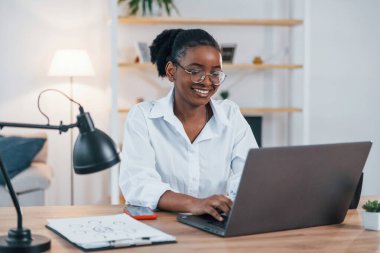 Dizüstü bilgisayarla çalışıyor. Beyaz gömlekli Afro-Amerikan kadın evde..