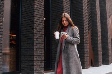 Gri ceketli genç kadın binanın yanında yürüyor ve elinde akıllı telefon ve bir bardak içki tutuyor..