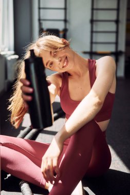 Yerde içkiyle otururken. Spor elbiseli genç bir kadın spor salonunda spor yapıyor..