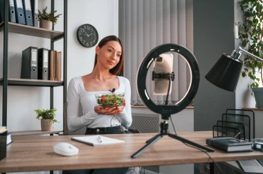 Beslenme uzmanı kamerada salata gösteriyor. Beyaz elbiseli genç ve güzel bayan ofiste..