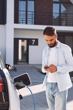 Şarj edilmeyi bekliyor, akıllı telefonu tutuyor. Genç şık adam, gündüz vakti elektrikli arabayla geziyor..