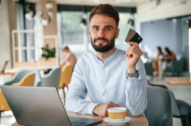 Kredi kartı tutuyor. Genç adam kafede oturuyor ve bilgisayarını kullanarak çalışıyor..