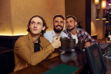 Oturuyor ve gülümsüyor. Üç arkadaş barda bira içip iyi vakit geçiriyorlar..